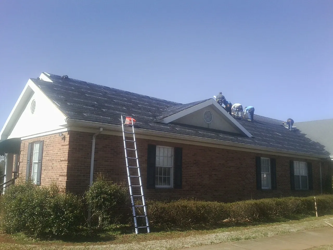Crew tearing shingles off a brick ranch roof with a ladder set — Emergency Roof Repair work by our crew in Farmington